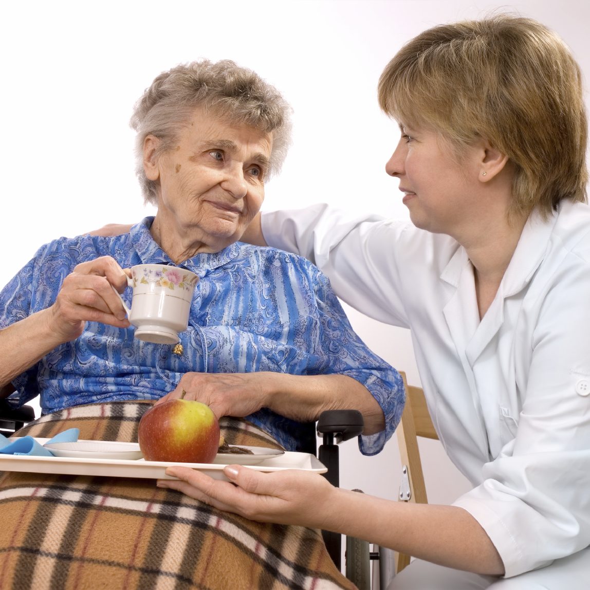 A caregiver offering an apple and a cup of tea to an elderly woman in a wheelchair.